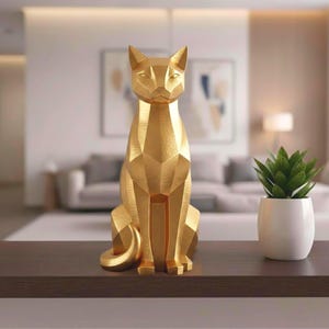 May include: A gold-colored geometric cat sculpture with a seated pose. The cat has a faceted design, giving it a modern look. The sculpture is on a dark brown surface, with a blurred background of a living room.