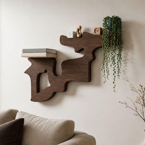 Può includere: Una mensola da parete in legno marrone scuro dal design astratto unico. La mensola contiene libri, un piccolo orologio, una statuina dorata e una pianta in vaso con viti verdi pendenti. Montata su una parete bianca.