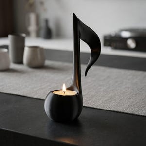 May include: A black candle holder shaped like a musical eighth note. The candle holder has a round base and a tall, curved stem with a flame burning inside. The background is blurred, with neutral tones.