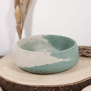 May include: A round, handcrafted concrete bowl with a two-tone design. The lower half is a light beige, while the upper half is a muted teal. The bowl sits on a natural wood slice, with dried ornamental grasses in the background.