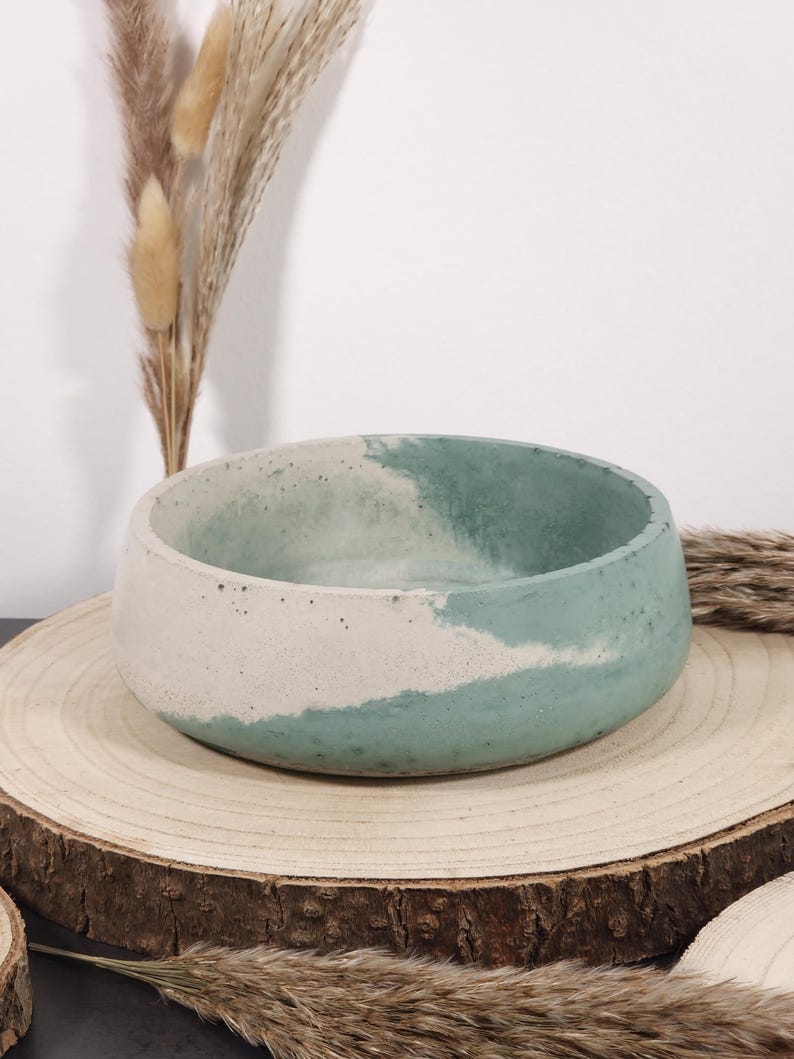 May include: A round, handcrafted concrete bowl with a two-tone design. The lower half is a light beige, while the upper half is a muted teal. The bowl sits on a natural wood slice, with dried ornamental grasses in the background.