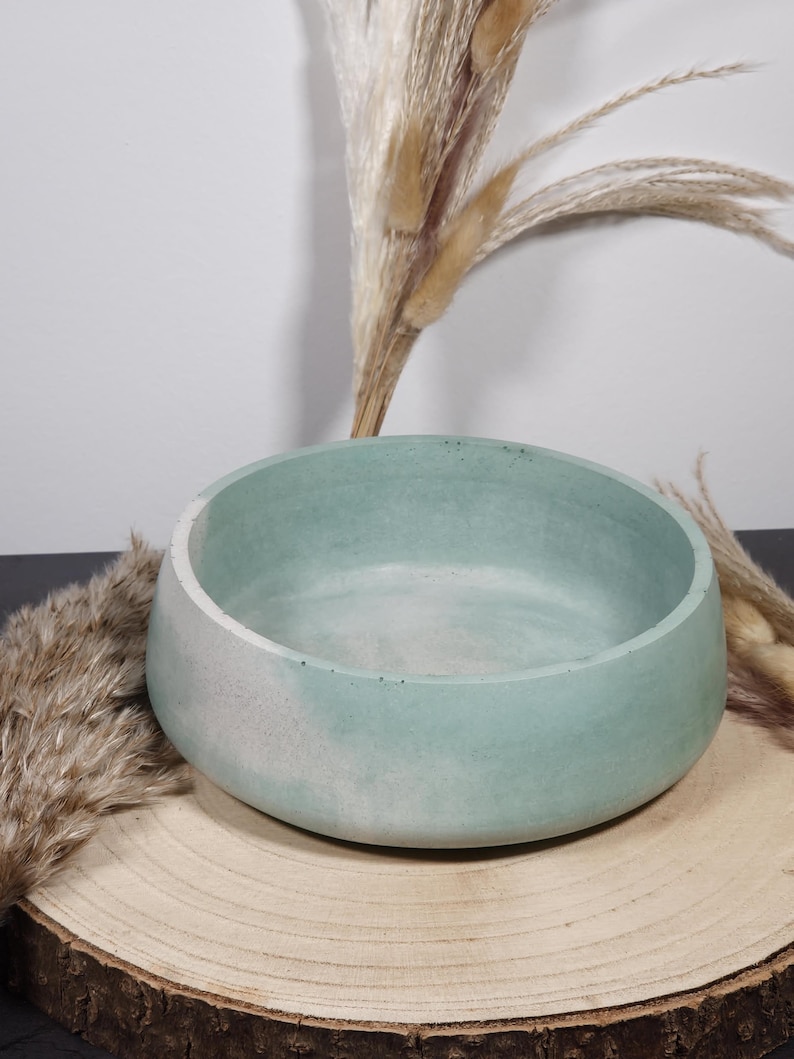 May include: A round, light blue concrete bowl with a white rim, set on a wooden slice. The bowl has a smooth interior and a textured exterior. Dried ornamental grass is in the background.