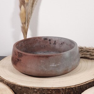 May include: A round, low-profile concrete bowl with a reddish-brown and gray color scheme. The bowl sits on a natural wood slice, with dried ornamental grasses in the background. The bowl's design is simple and modern, suitable for home decor.