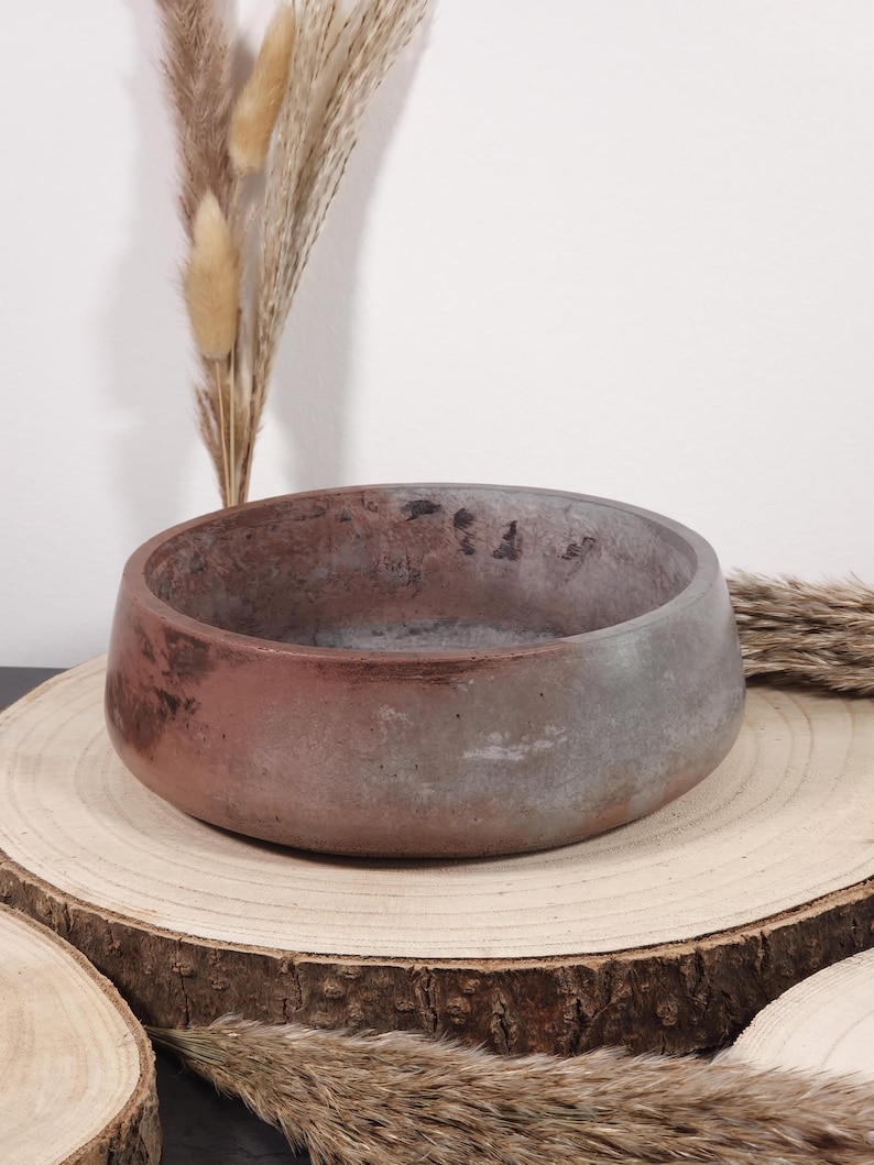 May include: A round, low-profile concrete bowl with a reddish-brown and gray color scheme. The bowl sits on a natural wood slice, with dried ornamental grasses in the background. The bowl's design is simple and modern, suitable for home decor.