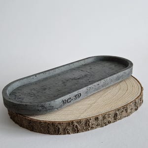 Könnte beinhalten: Eine ovale Schale aus grauem Beton mit strukturierter Oberfläche. Die Schale liegt auf einer Holzscheibe mit natürlichem Rindenrand. Der Text "DC-3D" ist auf der Seite der Schale sichtbar.