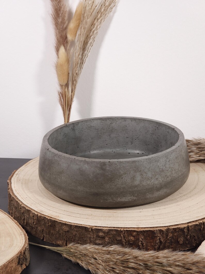 May include: A gray concrete bowl with a rounded shape, displayed on a natural wood slice. The bowl has a smooth, matte finish. Dried ornamental grasses are arranged near the bowl, adding a natural aesthetic.