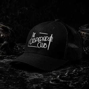Może przedstawiać: Czarna czapka typu trucker z siateczkowym tyłem i pełnym przednim panelem. Z przodu znajduje się biały haft z napisem THE DESPERADO CLUB oraz stylizowana grafika miecza wkomponowana w projekt, umieszczona pionowo po prawej stronie.