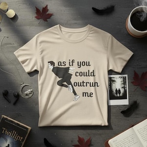Op de afbeelding: Beige t-shirt met de tekst "as if you could outrun me" en een zwart-witte afbeelding van een rennende persoon. Het shirt ligt op een donkere ondergrond met herfstbladeren, een boek en een kopje koffie.