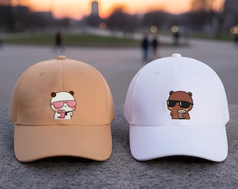 May include: Two baseball caps, one tan and one white, each with a cartoon bear graphic. The tan cap features a bear wearing sunglasses and holding a cup. The white cap has a bear wearing sunglasses. Both caps have a curved brim.