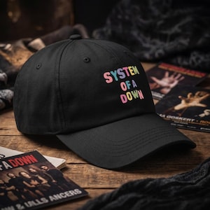 May include: Black baseball cap with the text "SYSTEM OF A DOWN" in colorful, pastel lettering. The cap is on a wooden surface with album art in the background. The hat has a curved brim and a button on top.