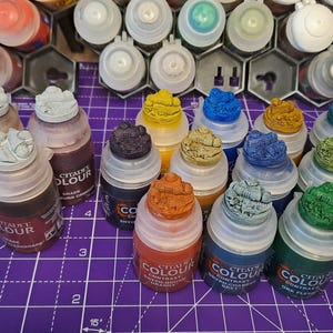 Könnte beinhalten: Eine Sammlung von Citadel Contrast Farben in verschiedenen Farben, darunter Blau, Grün, Braun und Orange. Die Farbflaschen sind in einem Raster auf einer violetten Oberfläche angeordnet. Jede Flasche hat eine kleine, detaillierte Miniaturfigur auf dem Deckel.