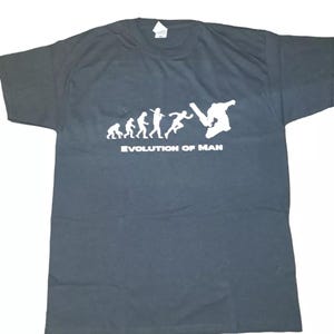 Könnte beinhalten: Schwarzes T-Shirt mit einer weißen Grafik, die die Evolution des Menschen darstellt und mit einem Skateboarder endet. Der Text "EVOLUTION OF MAN" ist darunter gedruckt. Das T-Shirt ist aus einem weichen Material.