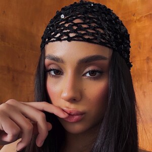 May include: A close-up portrait of a person wearing a black sequined head covering. The head covering is made of a net-like material and is adorned with small, shiny sequins. The person has dark hair and brown eyes.