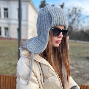 May include: A grey knitted hat with ear flaps and a long tie. The hat has a ribbed texture and is being worn by a person with long brown hair and sunglasses.