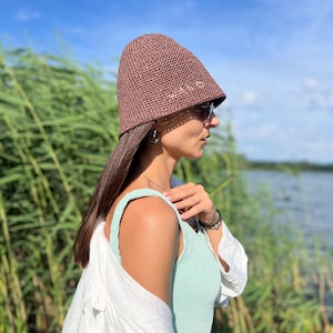 Op de afbeelding: Een vrouw draagt een bruine gehaakte zonnehoed met het woord "WILD" geborduurd aan de zijkant, een lichtgroen tanktop en een wit shirt. Ze staat in de buurt van een waterpartij met groene riet in de achtergrond.