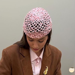 May include: A woman wearing a pink crocheted hat with sequins. She is wearing a brown blazer and a white button-down shirt.