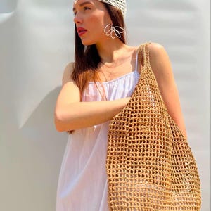 May include: A woman wearing a white crochet hat and a white dress is holding a brown crocheted tote bag.
