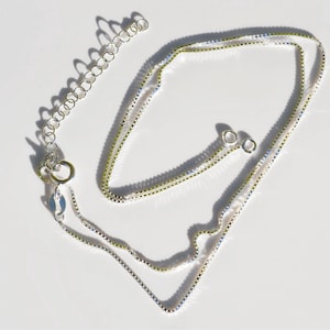 May include: A silver-colored box chain necklace featuring a delicate square link design. The necklace includes a circular spring ring clasp and an attached extender chain with several round links for adjustable length, reflecting light.