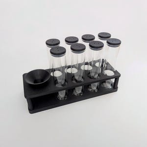 May include: A black wooden test tube rack holding eight clear glass tubes with black lids. A small black bowl sits on the left side of the rack. The rack is on a white background.