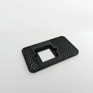 Puede incluir: Un soporte de teléfono rectangular negro con un patrón de panal en la superficie. El soporte tiene un recorte cuadrado en el centro y una pequeña sección elevada en un lado. El soporte está hecho de plástico.