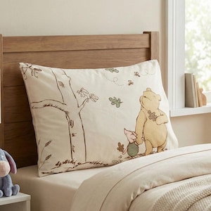 Pode incluir: Uma fronha bege com um desenho de Winnie the Pooh e Piglet. A ilustração mostra Pooh segurando um pote de mel e Piglet ao lado dele, com uma árvore e folhas. A fronha está em uma cama com um edredom bege.