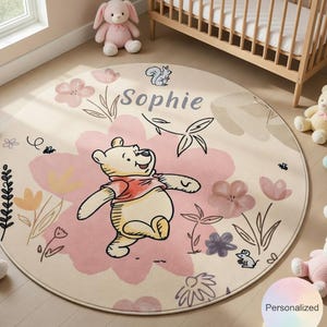Könnte beinhalten: Ein runder Winnie-the-Pooh-Teppich mit cremefarbenem Hintergrund und einem großen rosa Blumendesign. Der Teppich zeigt eine Cartoon-Illustration von Winnie the Pooh, florale Akzente und den Namen "Sophie".