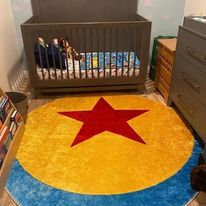 May include: A circular area rug featuring a large red star centered on a yellow background, surrounded by a blue border. The rug is positioned on a light-colored floor in front of a gray wooden crib and a gray dresser in a bedroom setting.