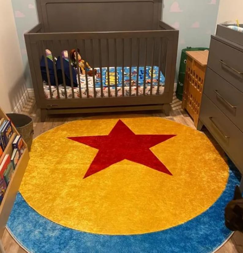 May include: A circular area rug featuring a large red star centered on a yellow background, surrounded by a blue border. The rug is positioned on a light-colored floor in front of a gray wooden crib and a gray dresser in a bedroom setting.
