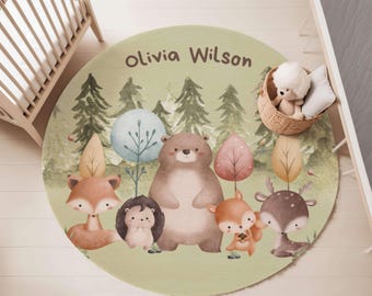 May include: A round area rug with a light green background featuring a watercolor forest scene with a bear, fox, hedgehog, squirrel, and deer. The name Olivia Wilson is printed in brown text at the top. A woven basket with a plush toy sits on the rug.