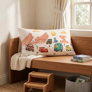 May include: A rectangular pillow featuring a printed desert landscape with cartoon characters from the movie Cars, including a red race car, a brown tow truck, a green van, and other vehicles, set against a backdrop of red rock formations and cacti.