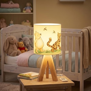 May include: A Winnie the Pooh themed lamp with a wooden tripod base. The lampshade features illustrations of Winnie the Pooh, a tiger, and dragonflies. The lamp is on a wooden nightstand with books and a remote control.