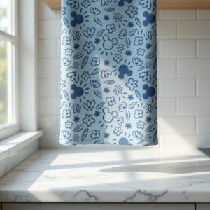 May include: A light blue kitchen towel with a repeating pattern of Mickey Mouse silhouettes, flowers, and leaves in a darker shade of blue. The towel is hanging in front of a white tile wall and a window.
