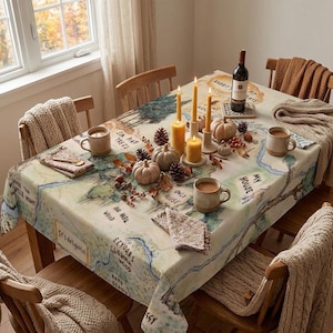 May include: A dining table set for a fall gathering. The table is covered with a map-themed tablecloth and decorated with pumpkins, candles, pinecones, and mugs. A bottle of wine sits on a coaster.