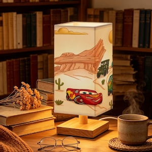 Puede incluir: Una lámpara de mesa rectangular con una pantalla de color claro que presenta una ilustración de dibujos animados de un coche de carreras rojo con el número 95, un vehículo militar verde y una furgoneta verde en un desierto con rocas y cactus.