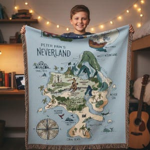 May include: A light blue woven throw blanket featuring a map of Neverland with the text "Peter Pan's Neverland." The map includes illustrations of Skull Rock, Misty Mountains, Mermaid Lagoon, and Pirate's Cove. The blanket has a fringed edge.