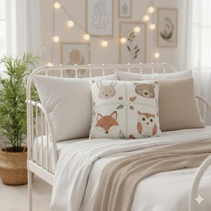 May include: A decorative pillow with a woodland animal design featuring a rabbit, bear, fox, and owl. The pillow is square and has a light beige background with a stitched grid pattern. Other pillows in white and beige are on a white metal bed frame.
