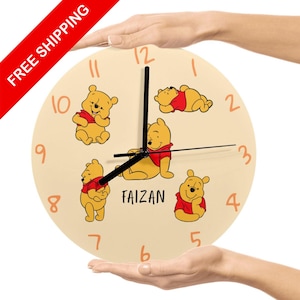 May include: A round wall clock with a cream-colored face featuring illustrations of a yellow bear in various poses, wearing a red shirt. The clock has black hands and orange numerals. The name "FAIZAN" is printed on the clock face.