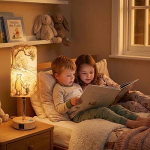 May include: A Winnie the Pooh themed bedside lamp with a cylindrical shade featuring an illustration of Pooh and Piglet. The lamp is on a wooden nightstand with two children reading a book in bed. Books and stuffed animals are on a shelf.