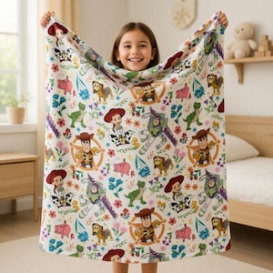 May include: A white blanket featuring a colorful pattern of Toy Story characters, including Woody, Buzz Lightyear, and Jessie, along with flowers and other elements. The blanket is held up by a person.