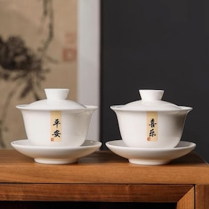 May include: Two white ceramic teacups with lids and matching saucers. The teacups have Chinese characters written on them. The teacups are on a wooden surface.