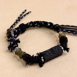 Könnte beinhalten: Ein handgefertigtes Armband mit einem schwarz-weiß geflochtenen Kordel. Das Armband hat schwarze und olivgrüne Perlen, silberfarbene Akzente und ein rechteckiges, geschnitztes schwarzes Holzmittelstück. Quasten sind an den Enden befestigt.