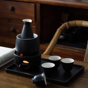 以下が含まれることがあります： 温める土台、ボトル、小さなカップ4つがついた黒い陶器の日本酒セット。セットは黒いトレイと木製のテーブルの上に置かれています。