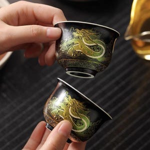 May include: Two black ceramic teacups with gold and green dragon designs. The cups are decorated with intricate patterns and are perfect for serving tea.