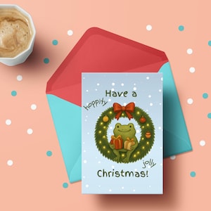 Może przedstawiać: Kartka świąteczna z żabą trzymającą prezenty w wieńcu, z napisem "Have a hoppity jolly Christmas!" na jasnoniebieskim tle. Karta jest w czerwonej kopercie z turkusowym wnętrzem, z filiżanką kawy na brzoskwiniowym tle.