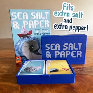 May include: A blue box set of the card game "Sea Salt & Paper" with the text "Fits extra salt and extra pepper!" The box includes a game box, a card holder, and cards. The game box features a paper boat design.