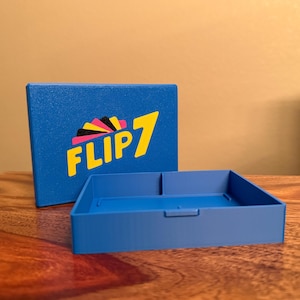 May include: A blue box and tray set with the text "FLIP 7" in yellow, black, and pink. The box is rectangular and the tray has two compartments. The set is on a wooden surface.