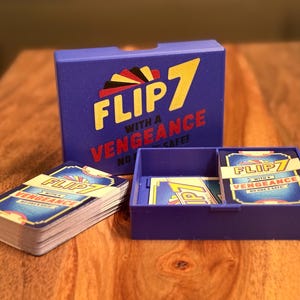 May include: A blue card game box and tray with the words "FLIP 7 WITH A VENGEANCE" in yellow and red. A stack of cards and a deck of cards are next to the box. The cards have a blue and gold design.