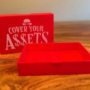 Op de afbeelding: Een helderrode doos en dienblad set. De doos heeft de witte tekst "COVER YOUR ASSETS" met een burger-icoon. De rechthoekige lade heeft een verdeeld interieur. De set staat op een houten oppervlak.