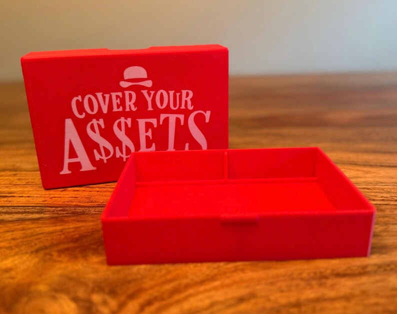 Op de afbeelding: Een helderrode doos en dienblad set. De doos heeft de witte tekst "COVER YOUR ASSETS" met een burger-icoon. De rechthoekige lade heeft een verdeeld interieur. De set staat op een houten oppervlak.