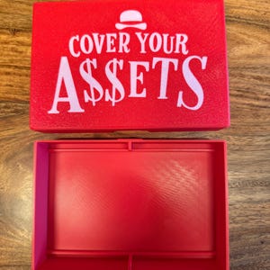 Op de afbeelding: Een rode rechthoekige doos met de tekst "COVER YOUR A$$ETS" in het wit. De doos is open en onthult een bijpassend rood interieur. Het ontwerp bevat een klein burger-pictogram boven het woord "COVER".